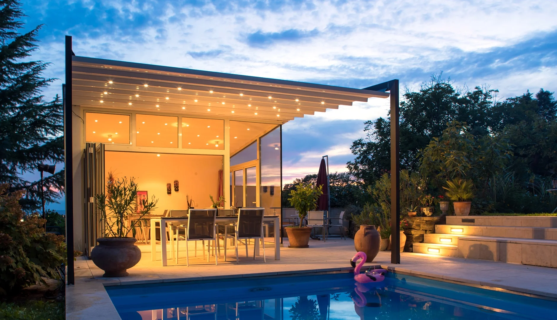 A modern outdoor patio with a swimming pool, illuminated by warm lighting, surrounded by plants and trees, with steps leading up to a seating area and a house with large glass windows and a ceiling with small string lights.