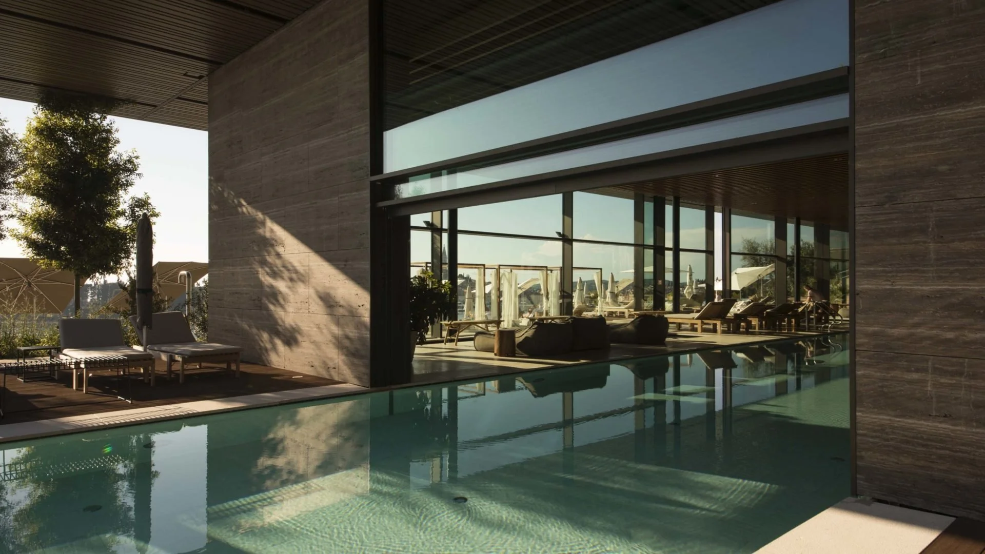 Modern indoor pool area with large glass windows overlooking a rooftop terrace with lounge chairs and umbrellas, sunlight casting shadows inside.