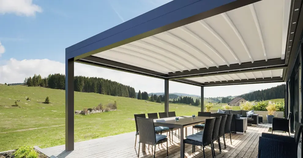 Outdoor patio with a dining table and chairs under a metal roof, overlooking a green hillside with trees and a cloudy sky.