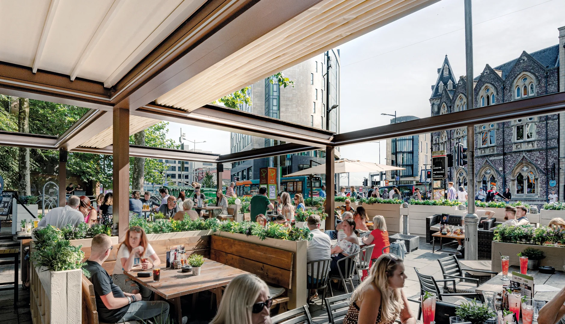 People dining at an outdoor restaurant patio with umbrellas and greenery in an urban setting with modern and historic buildings around.