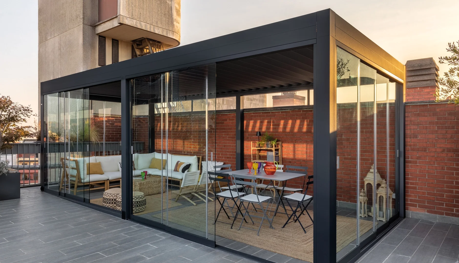 A modern rooftop terrace enclosed with glass sliding doors, featuring outdoor seating and dining areas, a brick wall, and city skyline views during sunset.