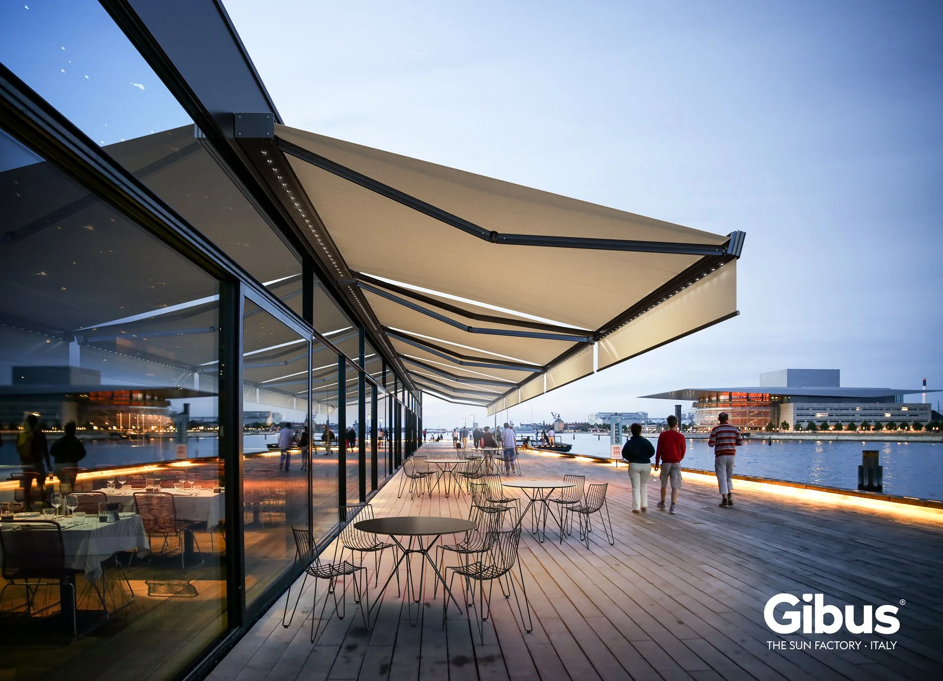 Outdoor dining area with round tables and wire chairs on a wooden deck, adjacent to a modern glass building with large windows. Overhead beige retractable awnings extend over the deck. The setting is near water with city buildings and a clear sky in the background at dusk.