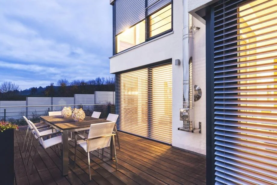 Modern outdoor patio with a wooden dining table and six white chairs, decorated with three large decorative objects, overlooking a fenced yard and trees during dusk, with illuminated windows and horizontal blinds on the house.