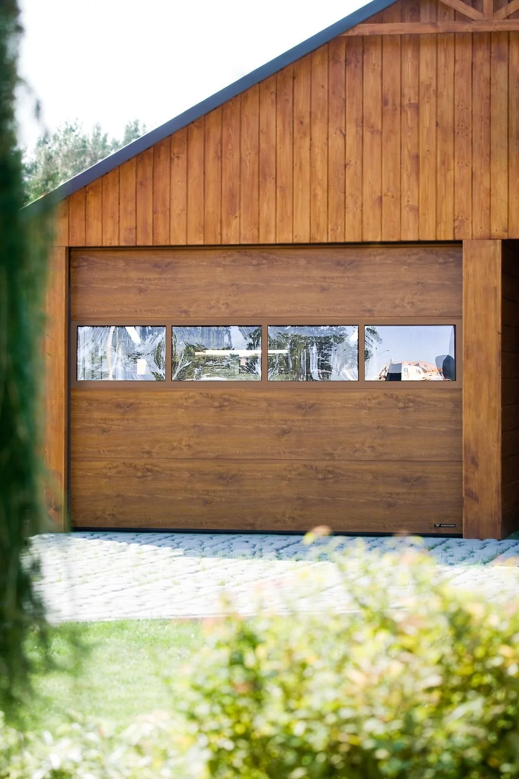A wooden garage door with a horizontal row of four windows, set in a wooden building with vertical siding, and surrounded by trees and bushes.
