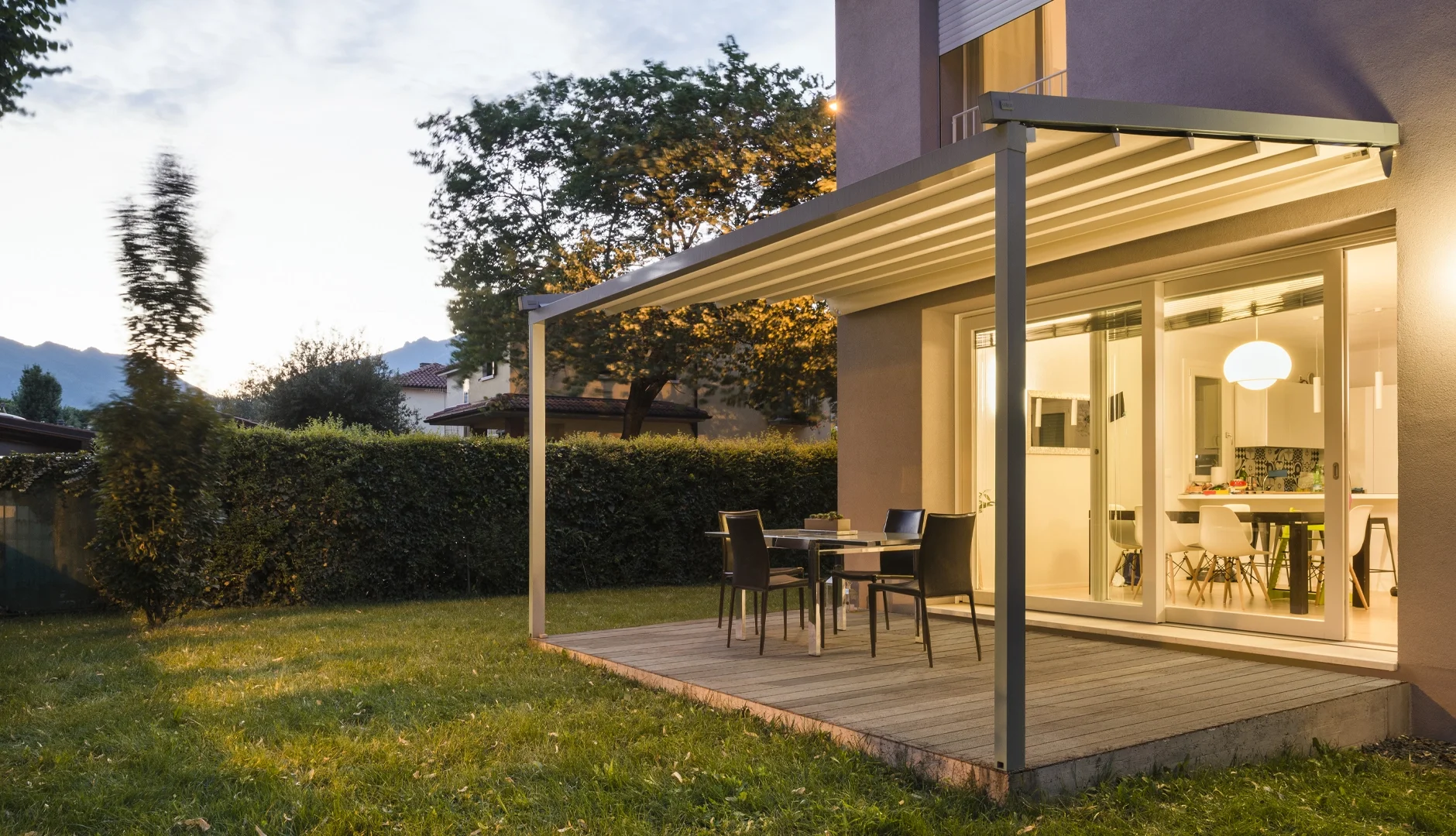 Backyard patio with outdoor dining table surrounded by four chairs, adjacent to a house with large sliding glass doors revealing a lit interior kitchen and dining area, during twilight.