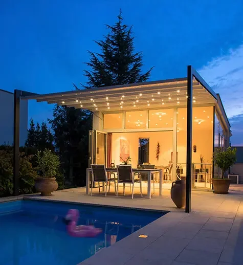 Modern outdoor patio with dining area, string lights, potted plants, beside a swimming pool at dusk