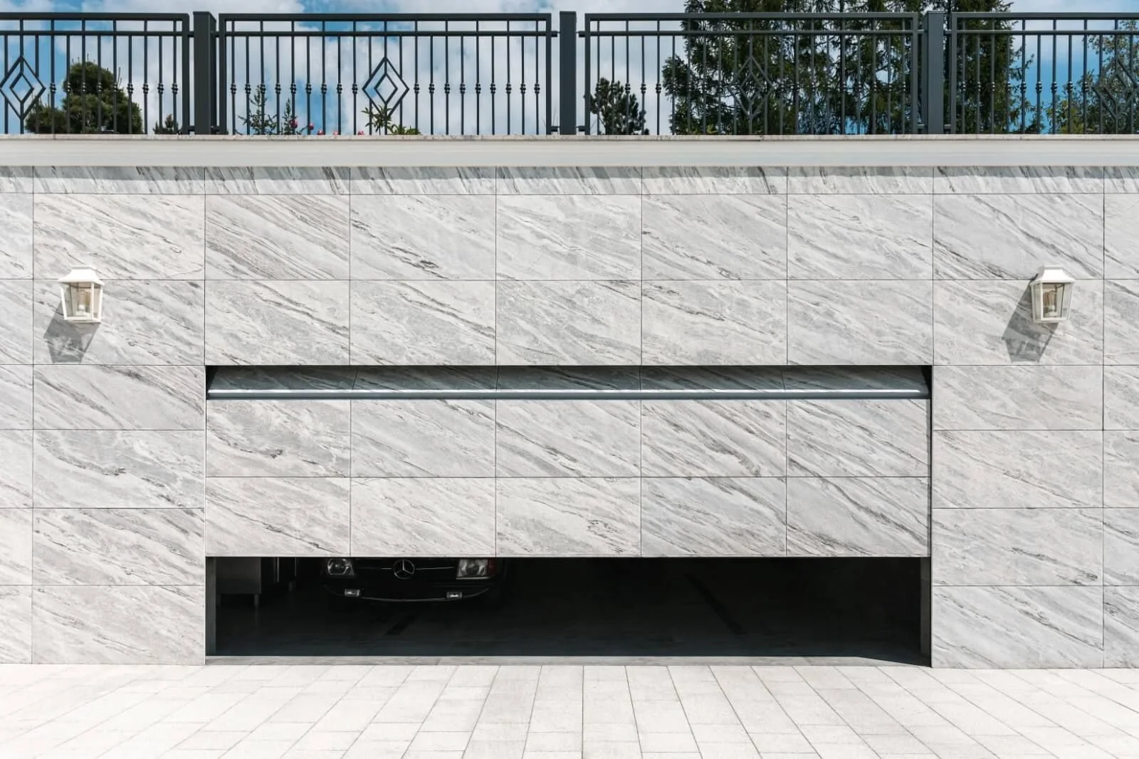 Exterior of a modern building with a garage door, marble-like wall, wall-mounted lights, and a black Mercedes-Benz car partially visible inside.