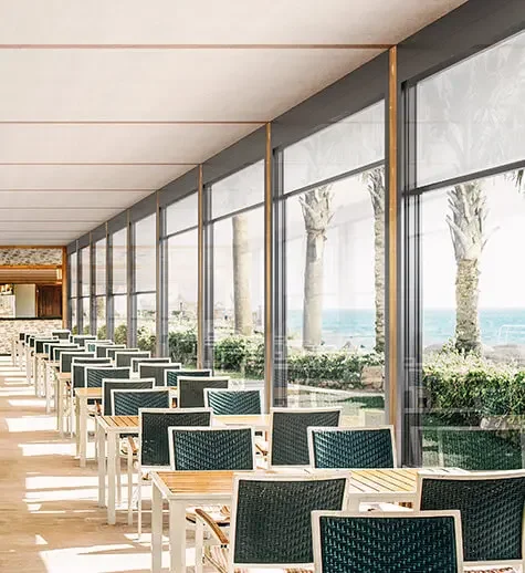 Indoor dining area with tables and chairs near large floor-to-ceiling windows overlooking palm trees and ocean.