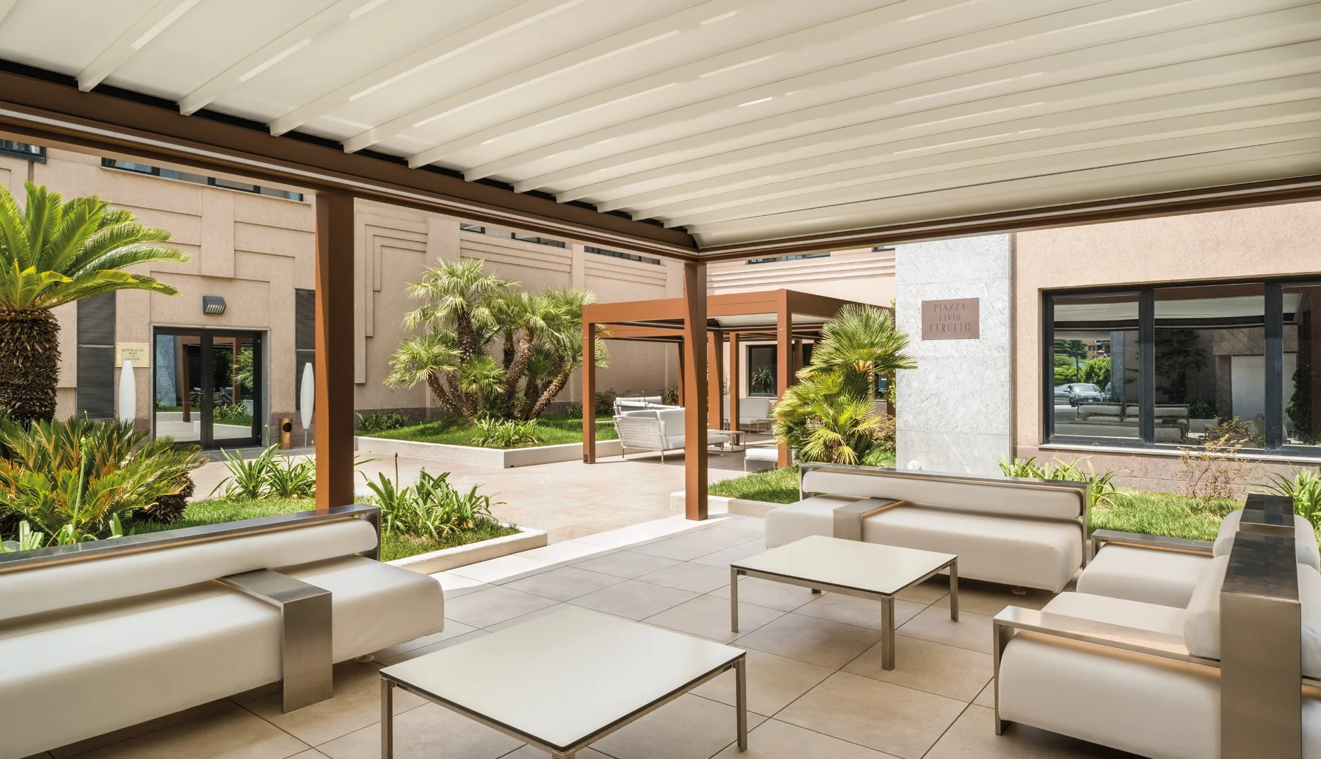 Outdoor seating area with white sofas and tables, surrounded by greenery including tall palm trees, covered by a white pergola in a modern building courtyard.