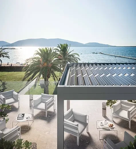 Outdoor patio with white chairs and tables, overlooking water and palm trees with a mountain range in the background.
