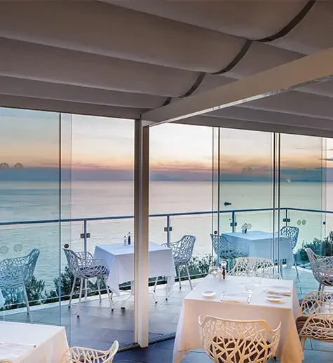Upscale restaurant with dining tables set for guests, overlooking ocean through large glass windows at sunset.