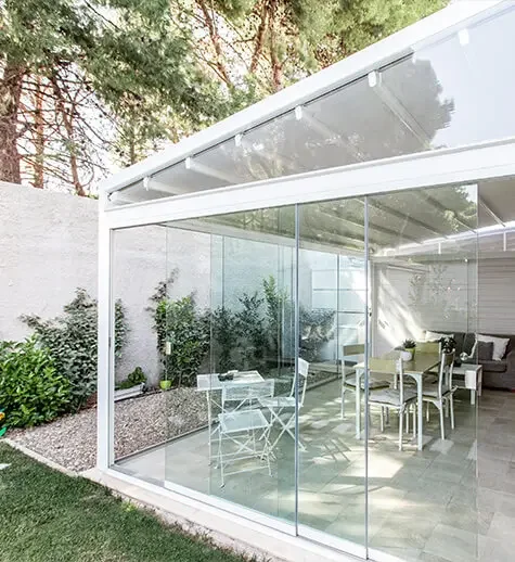 Modern glass-enclosed sunroom with outdoor garden view, containing a dining table with chairs, a sofa, and potted plants.