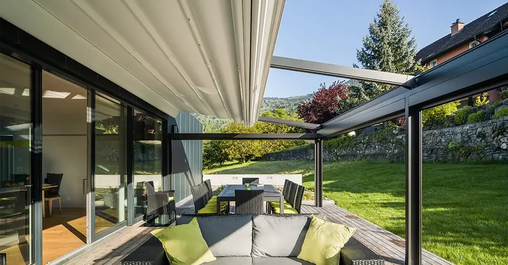 Outdoor patio area with a cushioned seating in front, a dining table with chairs in the middle, and glass sliding doors leading into a house on the left, surrounded by a green lawn and trees.
