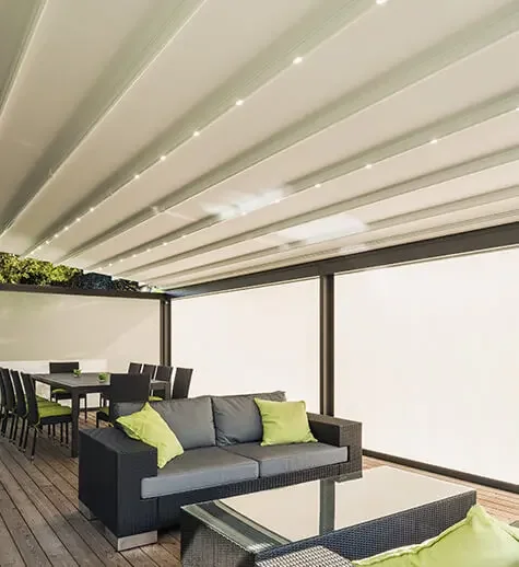 Outdoor patio with a modern black sofa, glass-topped coffee table, and six chairs at a long dining table under a white pergola with string lighting.