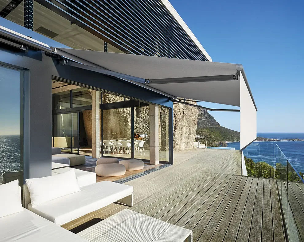 Modern rooftop terrace with white outdoor furniture, glass railing, and ocean view, featuring a rocky hillside in the background.
