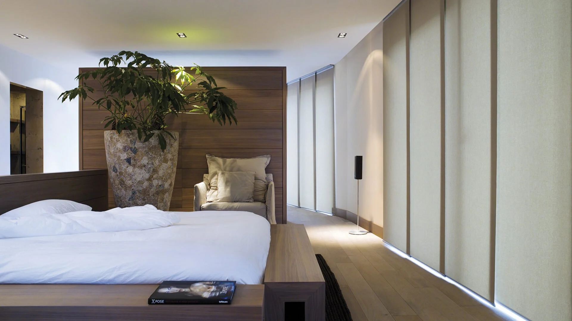 Modern bedroom with a large bed, white bedding, and a wooden headboard and side table. A large plant in a textured stone pot is behind the bed, with beige curtains on the right and a small armchair with pillows near the plant. Recessed ceiling lights and a floor lamp are visible.