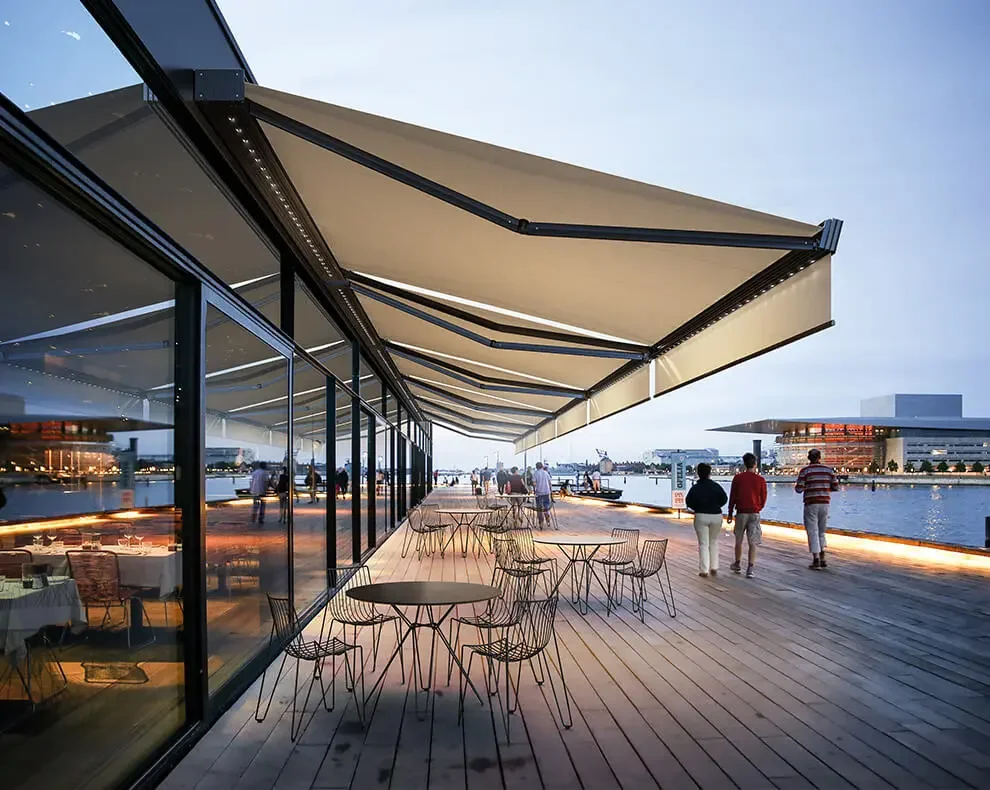 Outdoor restaurant or cafe patio with tables and chairs, glass windows, large beige awning, overlooking a waterway with modern buildings in the background, during dusk.