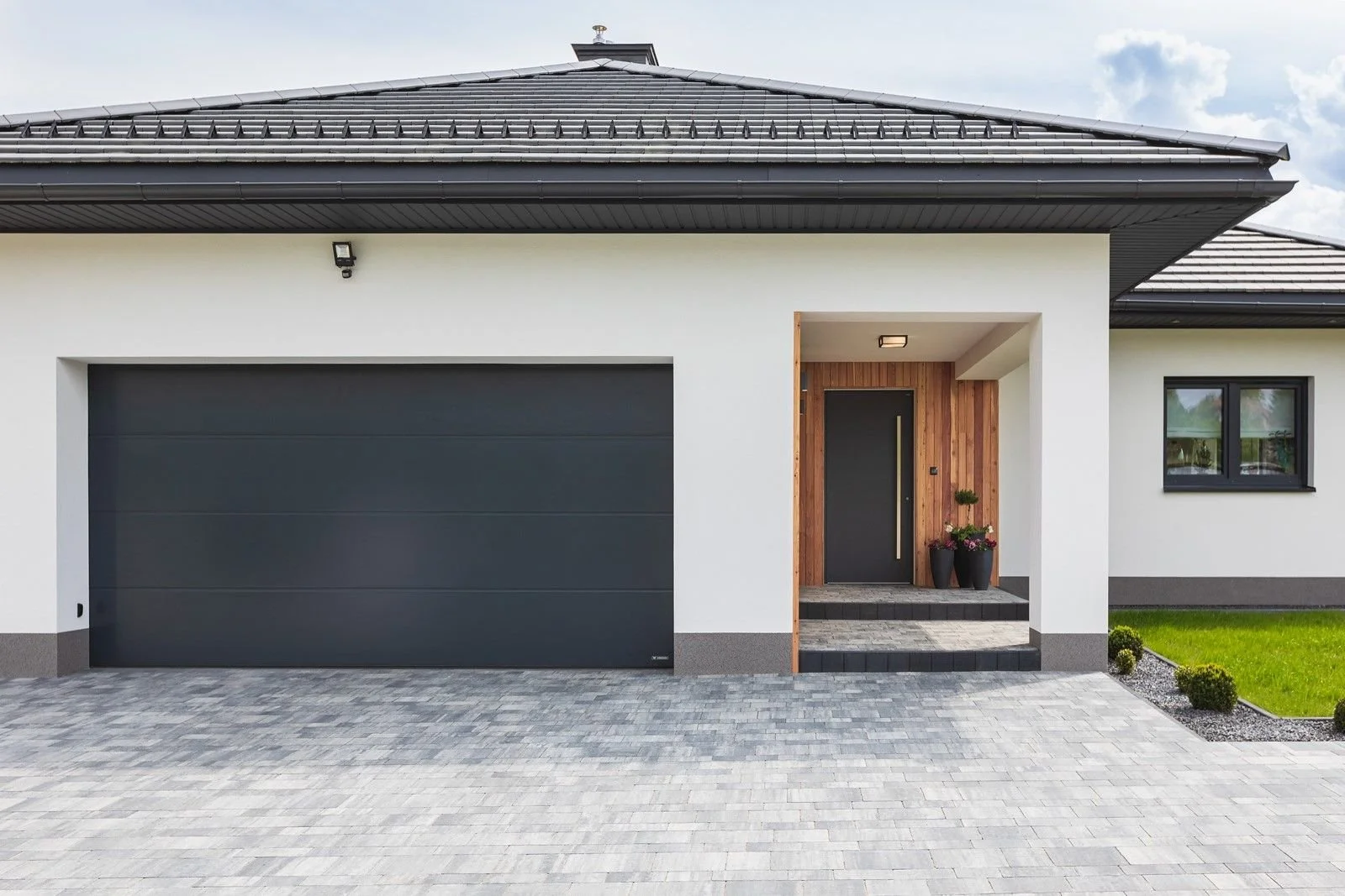 Modern house exterior with white walls, black garage door, and wooden front door, surrounded by a paved driveway and small landscaped bushes.