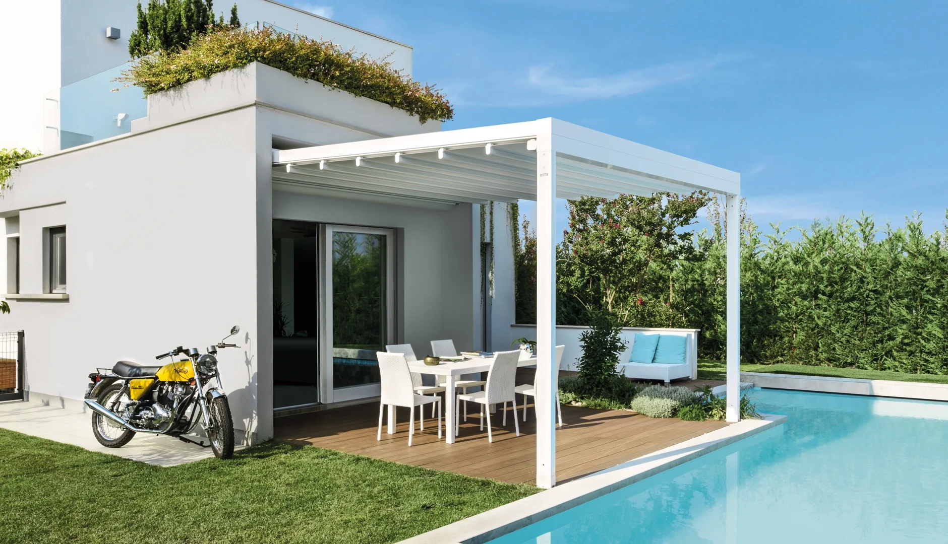 Modern backyard with a white house featuring a covered patio area with outdoor dining furniture, a yellow motorcycle parked outside, a swimming pool, and lush green landscaping.