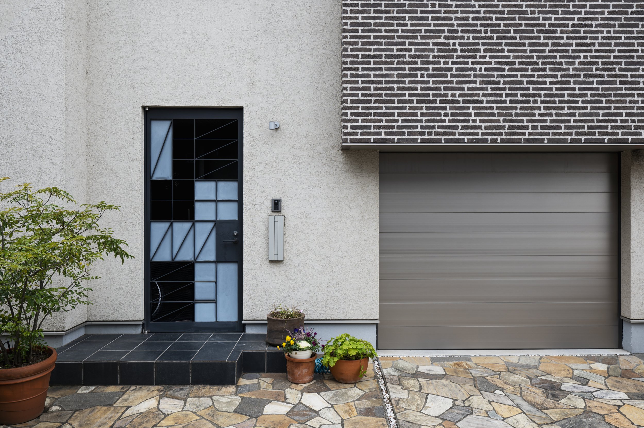 Modern house exterior with black and glass front door, gray garage door, potted plants, stone pathway, and textured beige wall with brick accents.