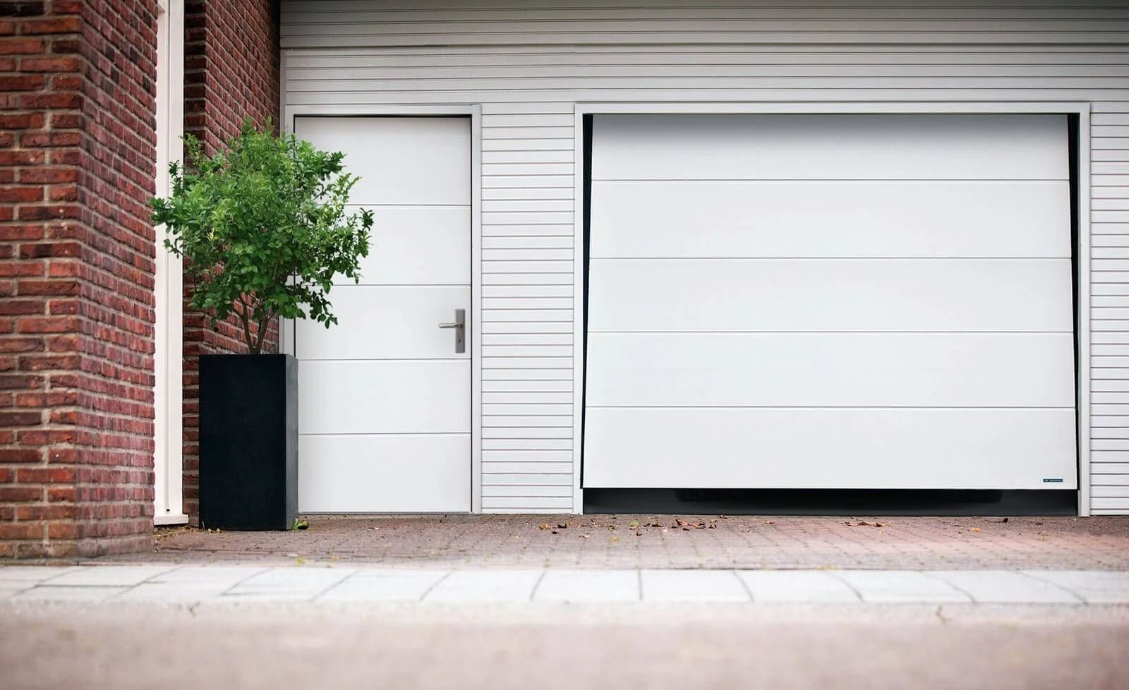 Front view of a garage with a large white sectional door and a smaller white side door, with a potted bush on the left side.