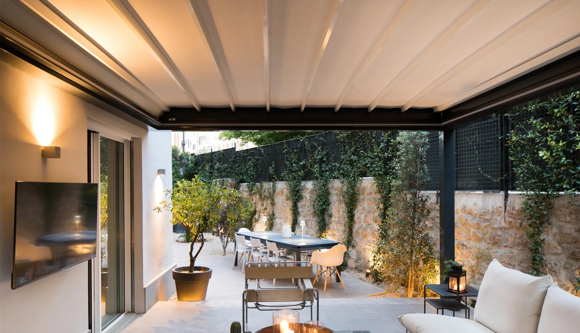 Modern outdoor patio with a covered roof, seating area, potted plants, string lights, and a stone wall with climbing plants.