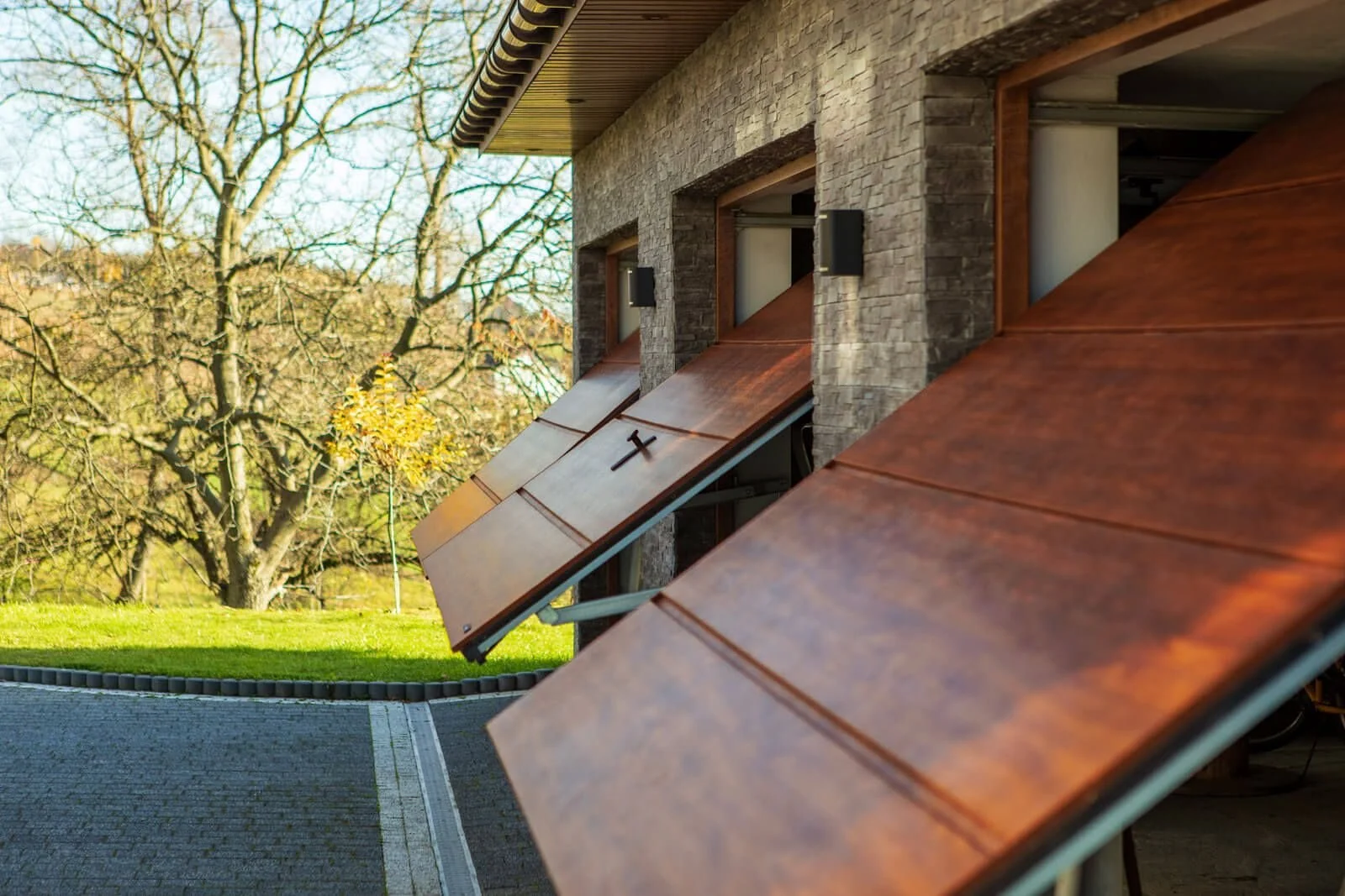 Three garage doors with wooden panels and small windows in a stone building, with leafless trees and a green lawn in the background.
