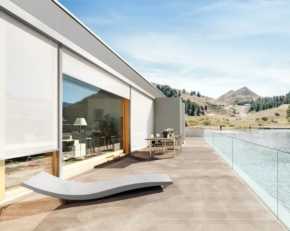 Modern house exterior with a spacious balcony overlooking a lake and mountain landscape, featuring a lounge chair and dining table with chairs.