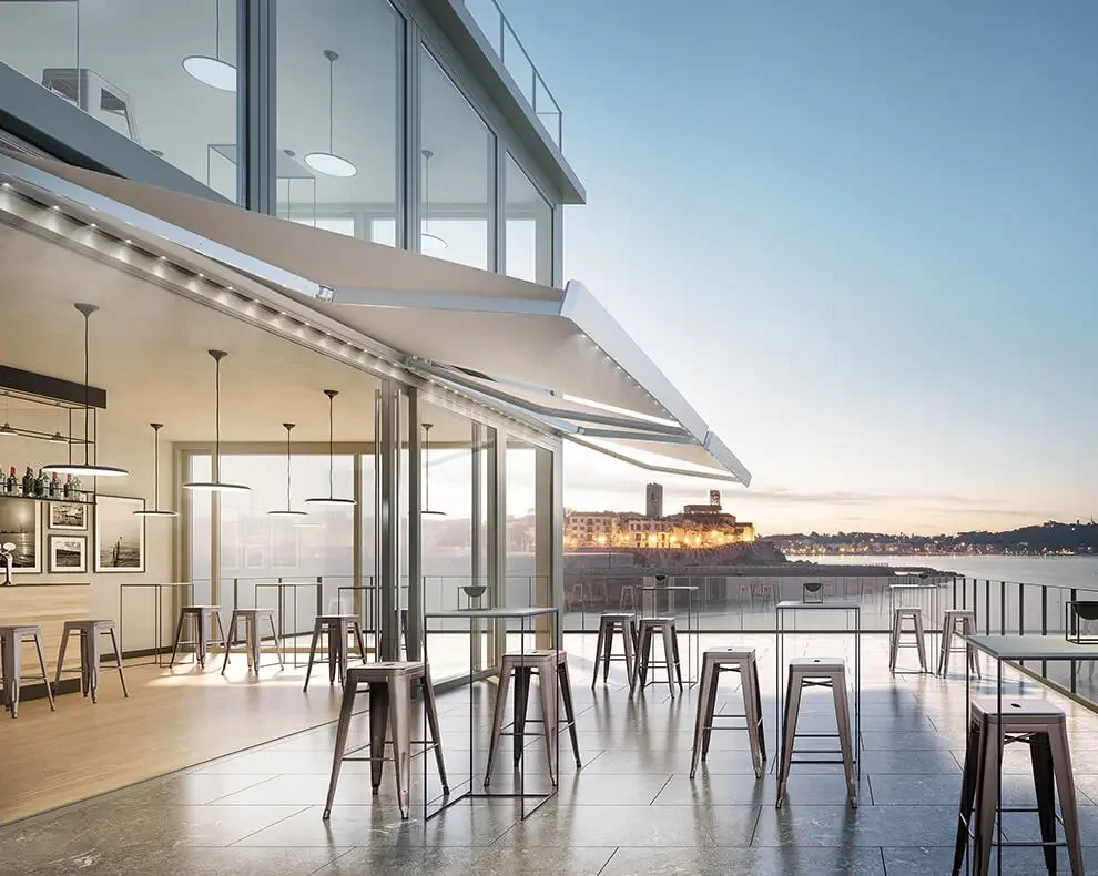 Modern outdoor terrace overlooking a body of water with city buildings in the distance, featuring bar-height tables and stools, glass railing, and a partly covered interior space with hanging lights.