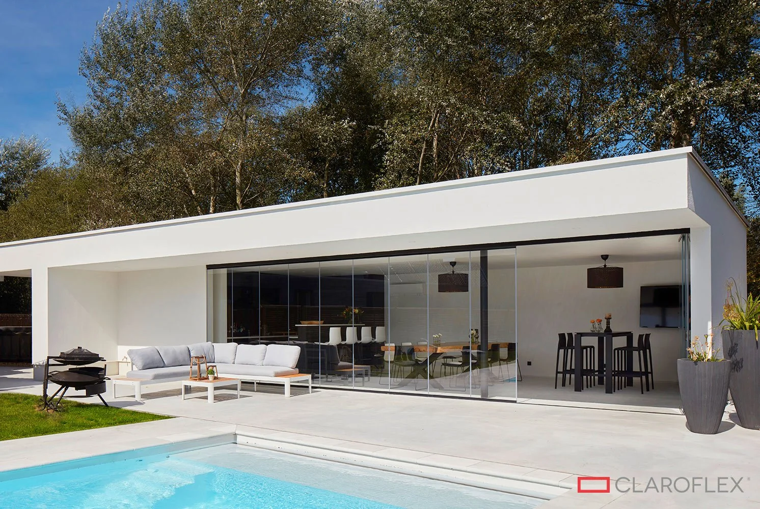 Modern house with large glass sliding doors, outdoor seating area, and swimming pool in the foreground, surrounded by trees.