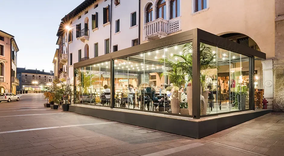 Glass-walled restaurant or café on a city street with potted plants and people dining inside during early evening.