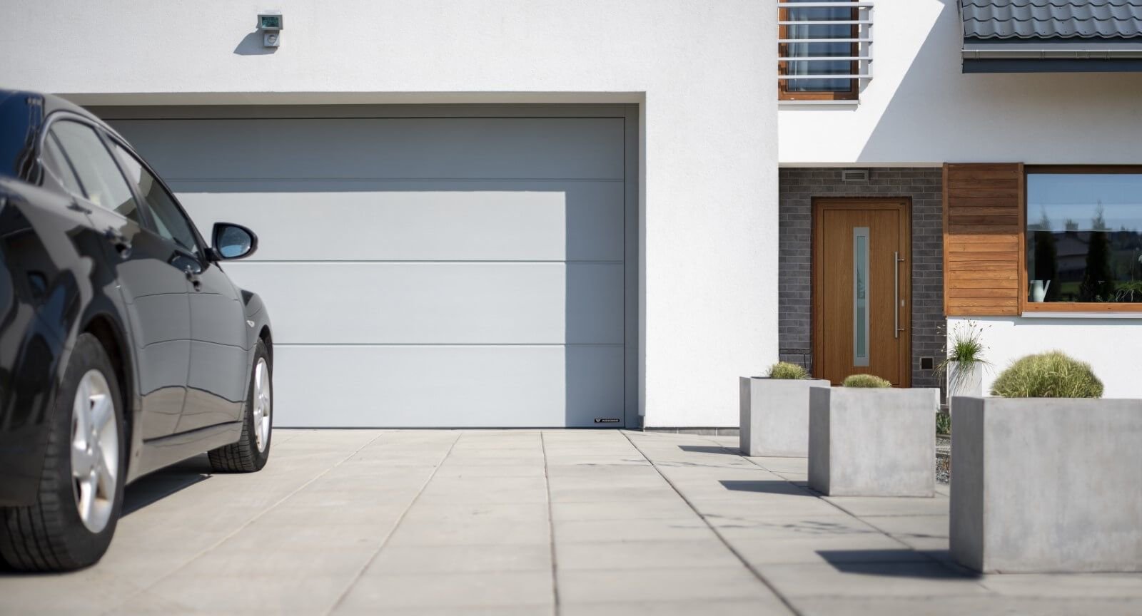 Modern house exterior with white walls, a gray garage door, a wooden front door, and a black car parked on the driveway. Decorative concrete planters with plants are in the yard.