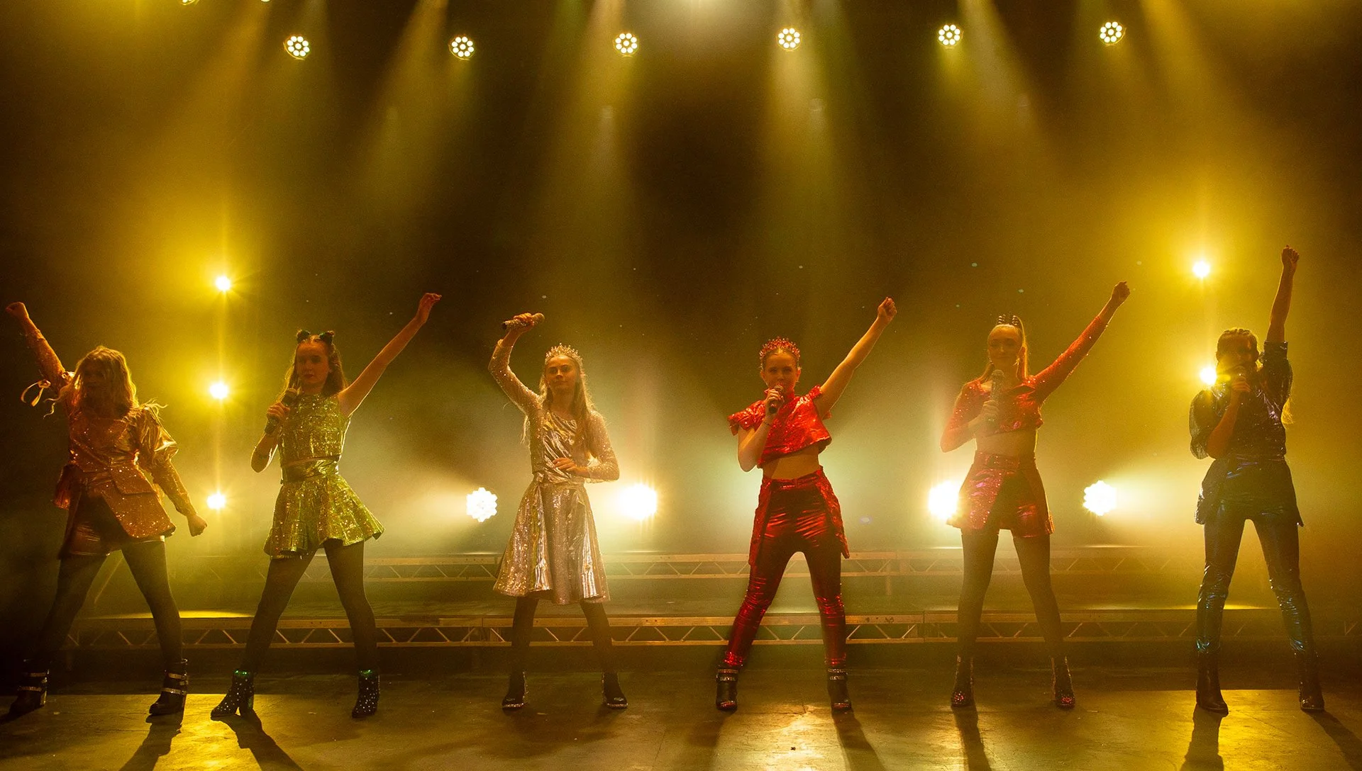 Six girls in colorful, shiny outfits perform on a stage with bright yellow lights shining behind them.