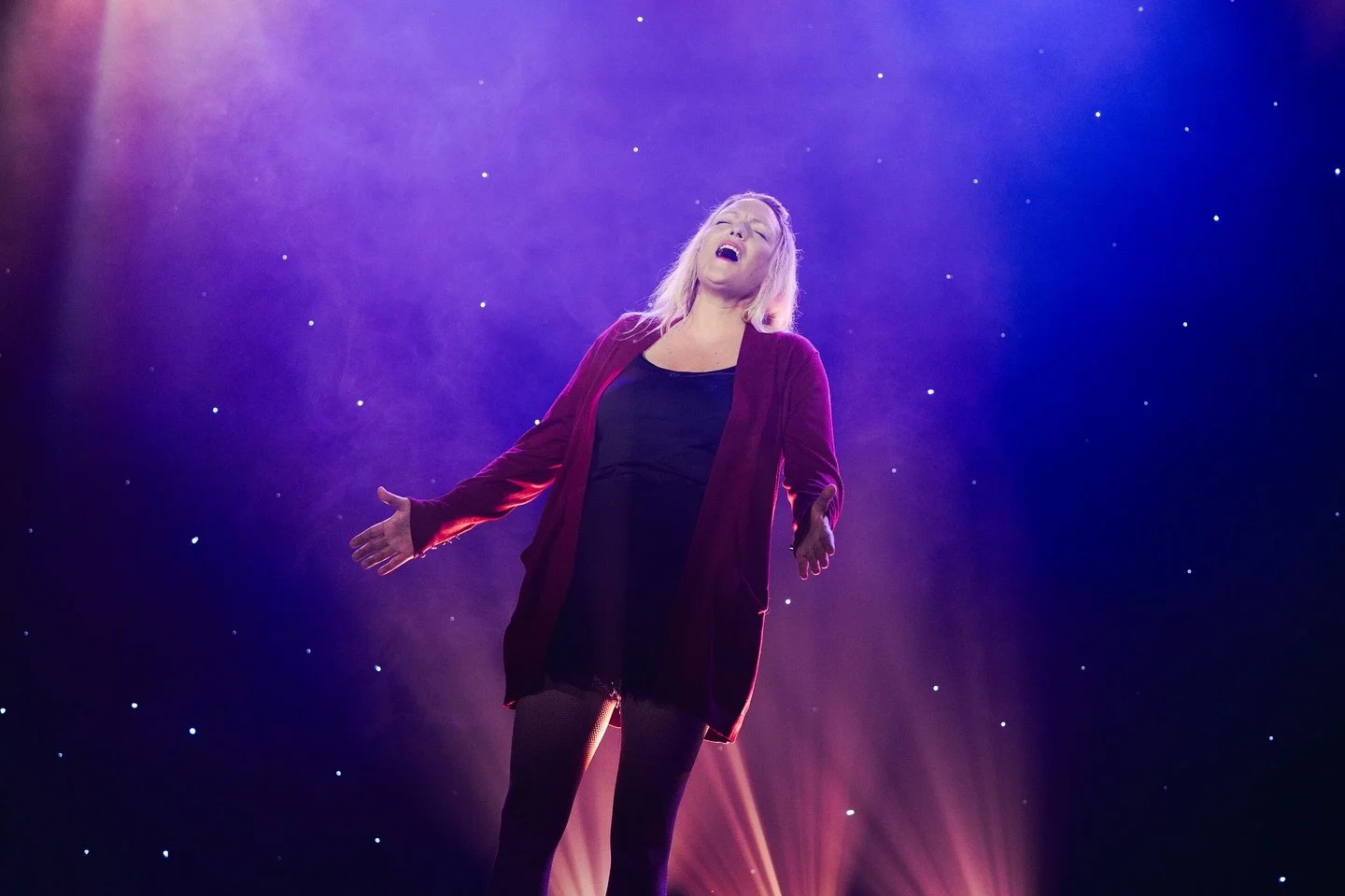 A woman singing passionately on stage with a purple and blue starry background, pink stage lights shining down.