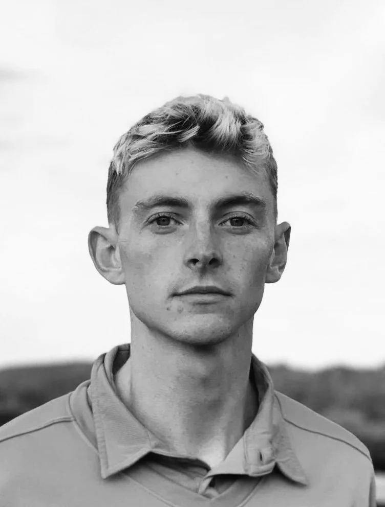 A young man with light-colored hair, wearing a collared shirt, standing outdoors in a natural landscape, looking at the camera.