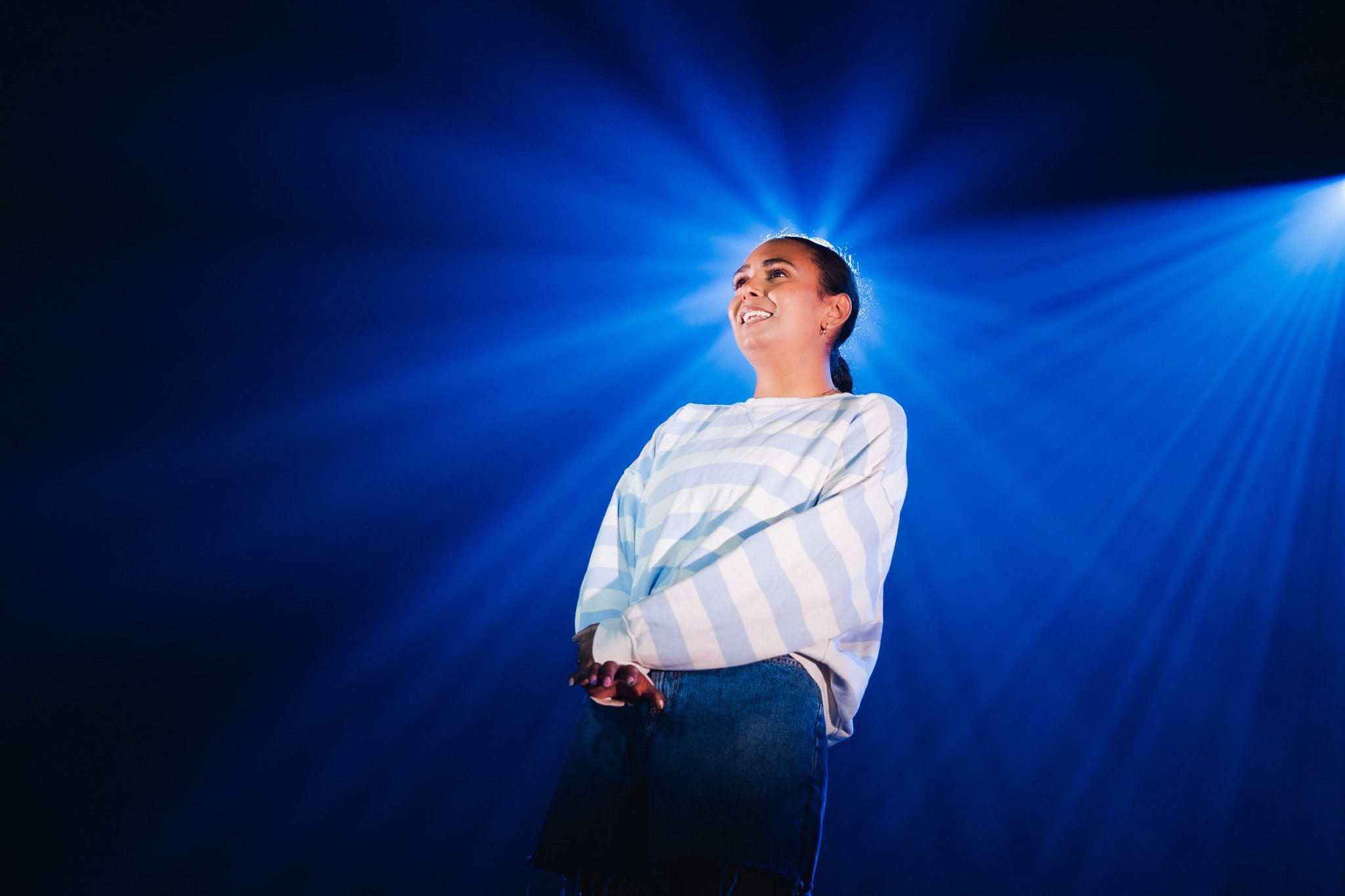 A young woman smiling confidently on stage, illuminated by dramatic blue backlighting.