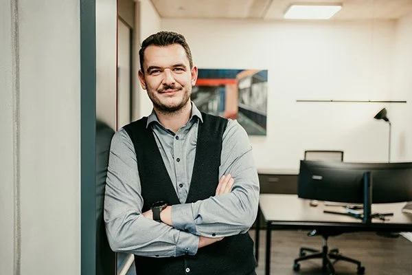 Ein Mann steht in einem modernen Büro mit weißen Wänden, einem Tisch, einem Computer und einer Stehlampe, und lächelt.