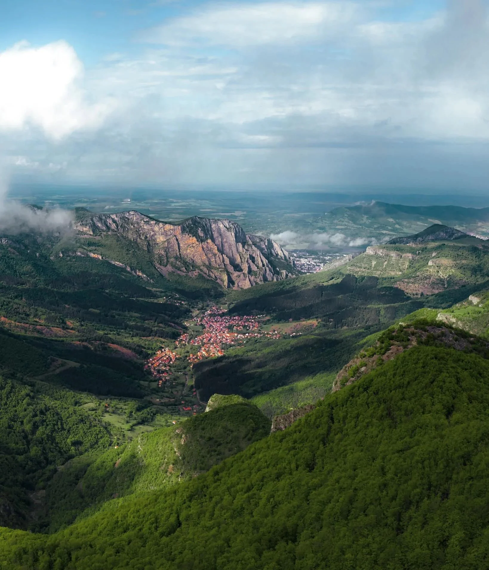 vue panoramique de montagnes verdoyantes, avec un petit village au centre et des nuages dispersés dans le ciel.