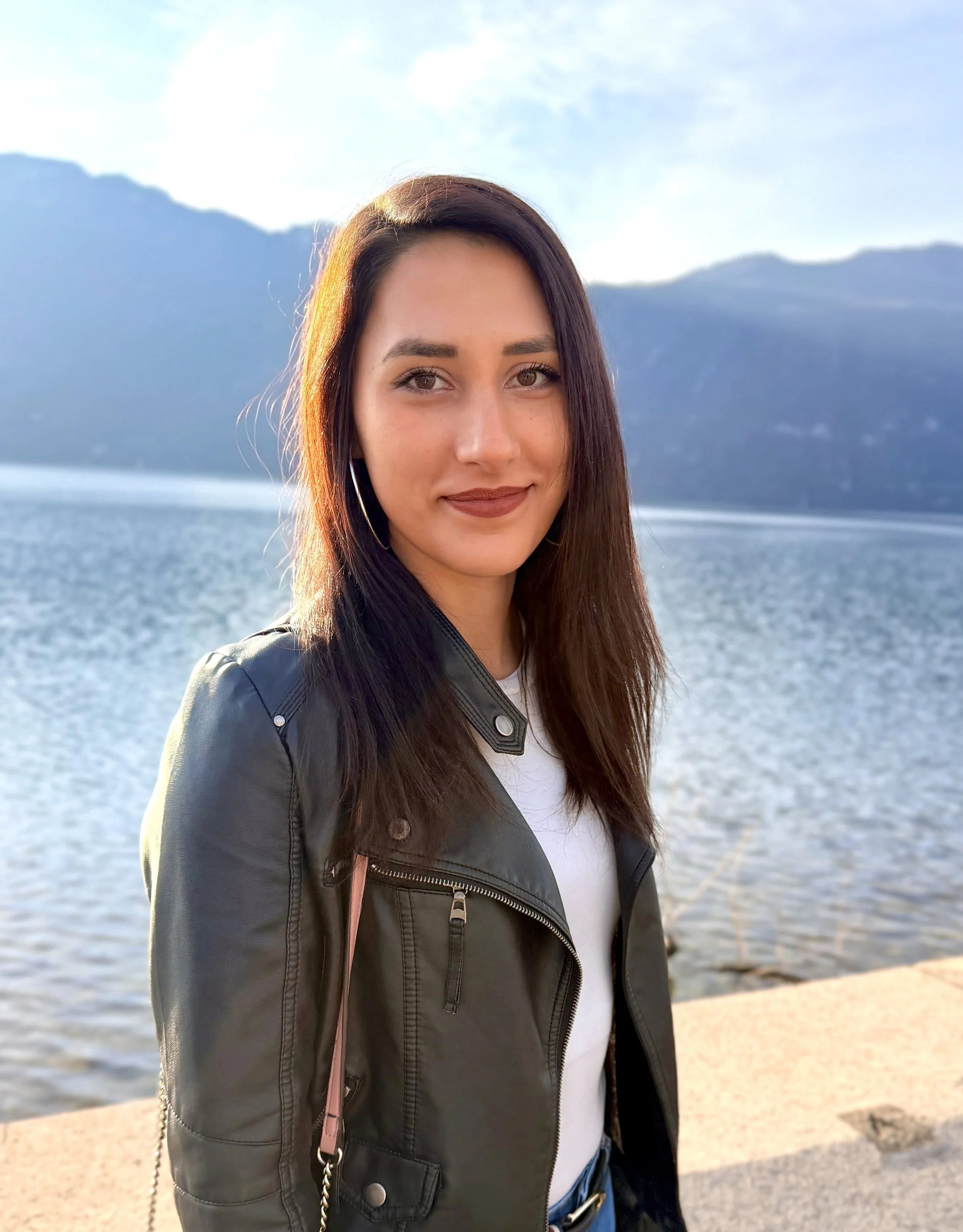 Jeune femme bulgare avec cheveux bruns foncés, souriante, portant un blouson en cuir noir, près d'un lac avec montagnes en arrière-plan, en fin d'après-midi ensoleillé.