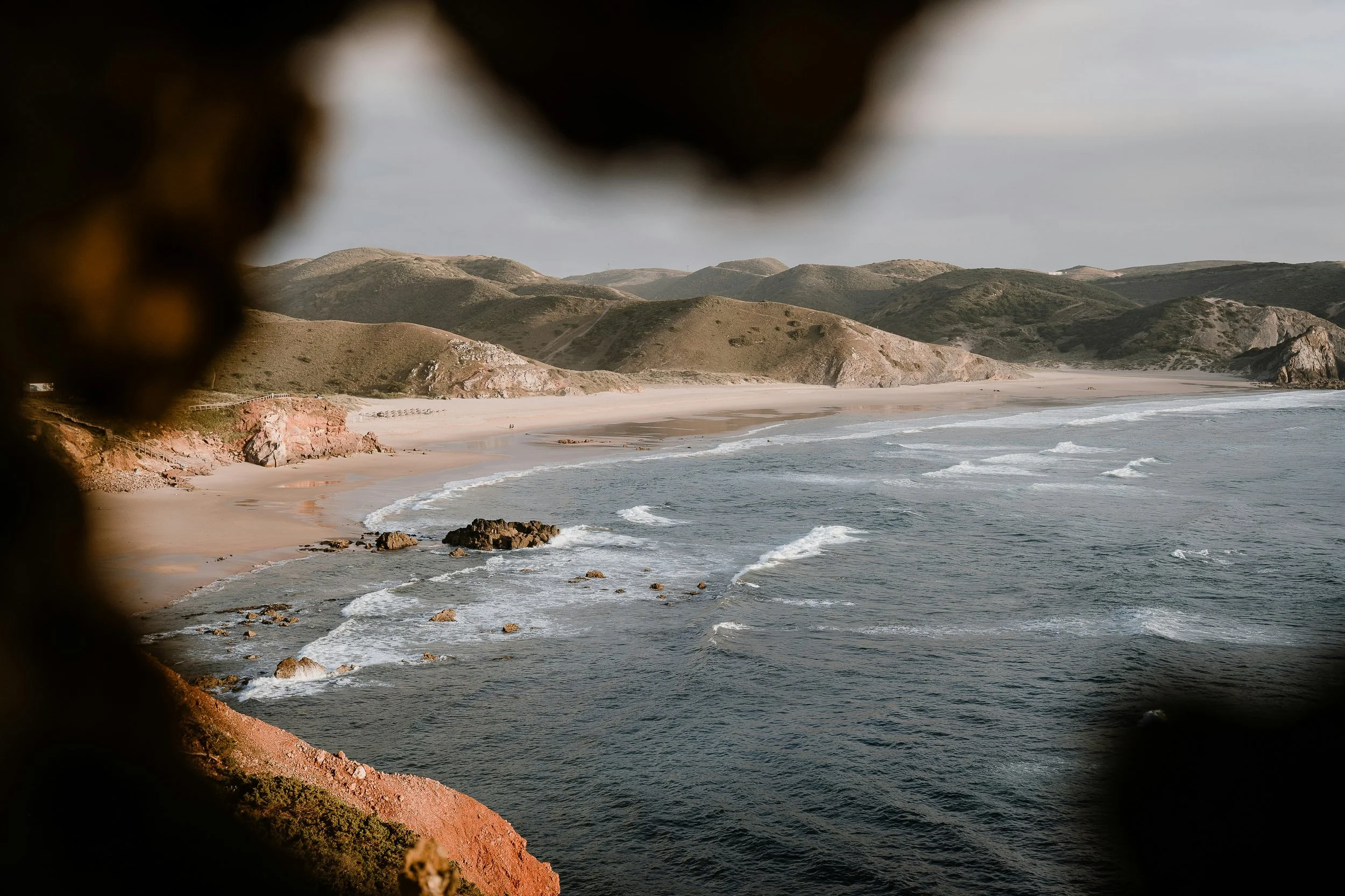 Vue de la plage de sable à travers une ouverture dans des rochers, avec des vagues et des montagnes en arrière-plan.