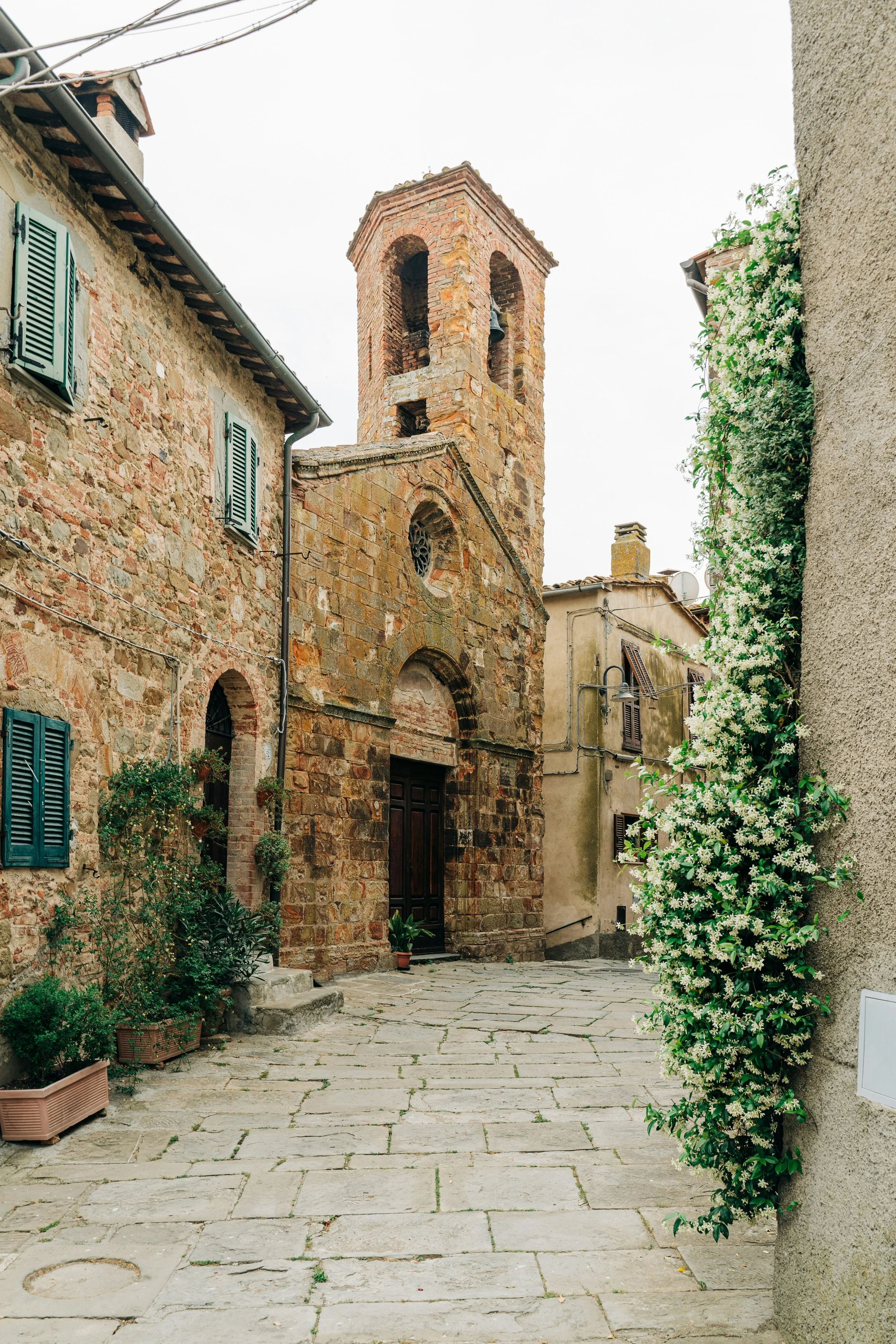 Une vieille église en pierre en Bulgarie avec un clocher, située dans une rue pavée entourée de bâtiments avec des volets en bois verts et des plantes fleurissant sur le côté.