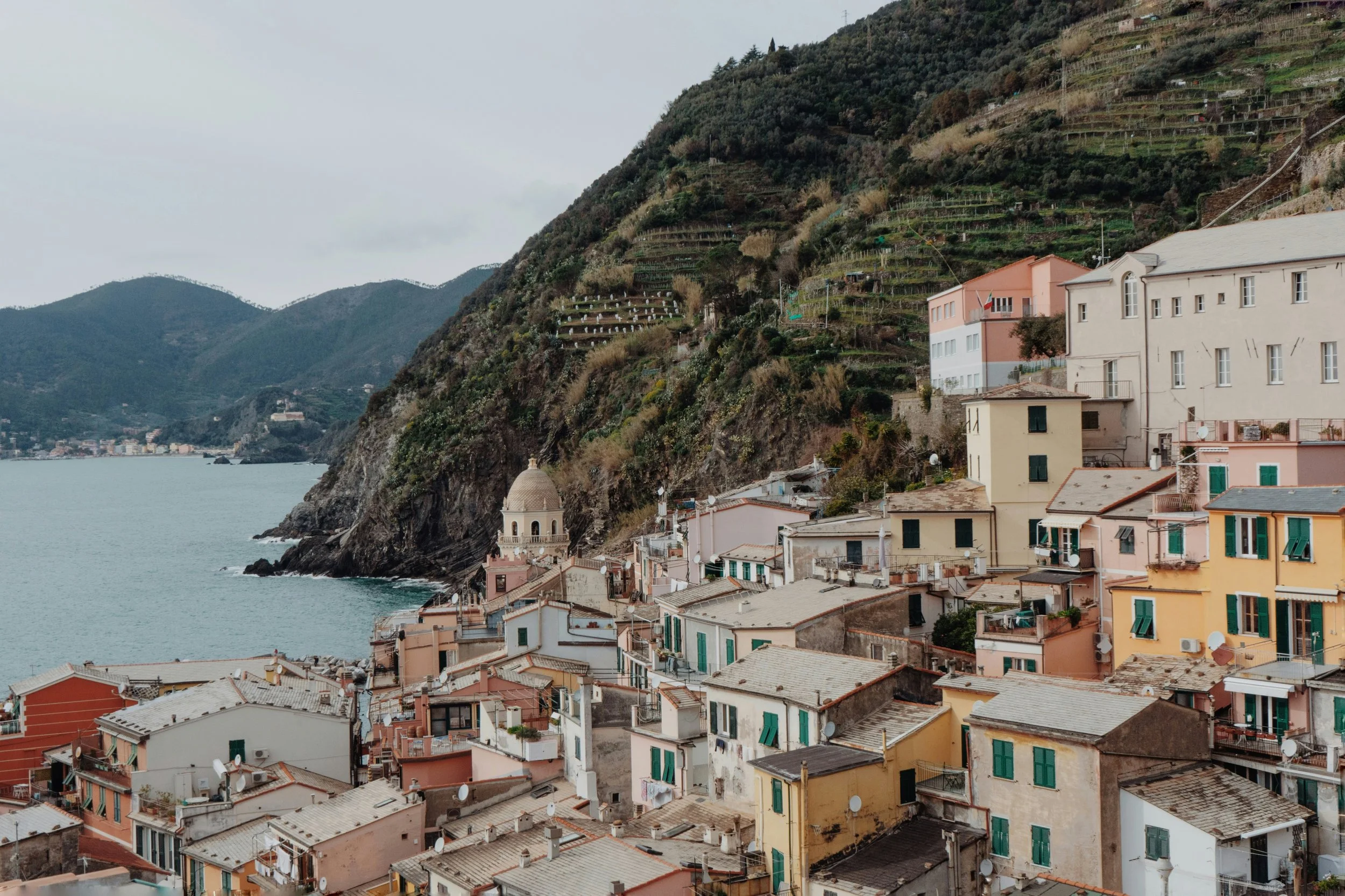 Villas colorées dans un village côtier, avec une église à dôme, sur une colline en pente, près d'une mer calme, sous un ciel nuageux.