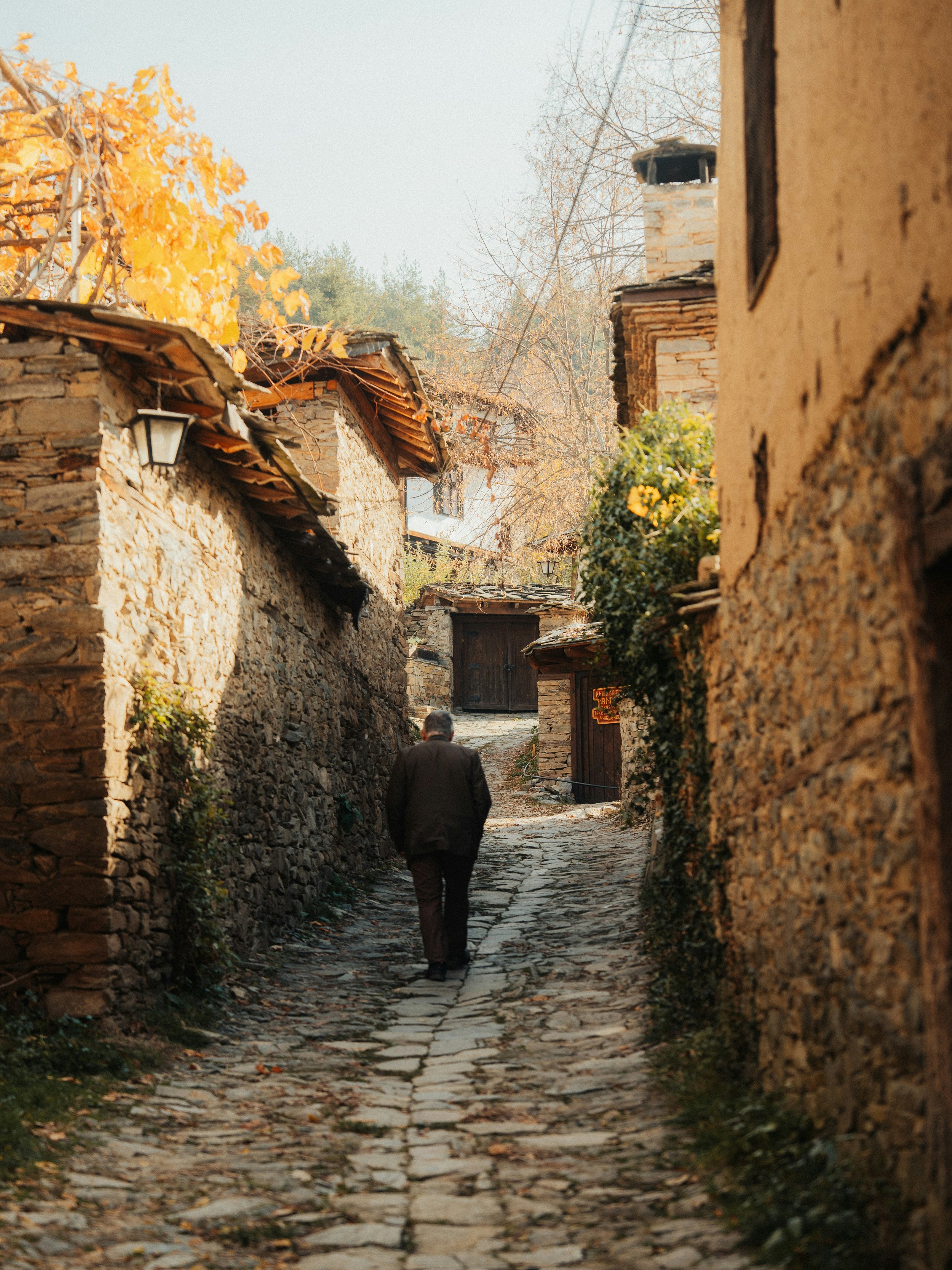 Un homme marche seul dans une ruelle pavée entre des maisons en pierre aux toits en tuiles, en automne, avec des arbres aux feuilles jaunes et brunes en arrière-plan.