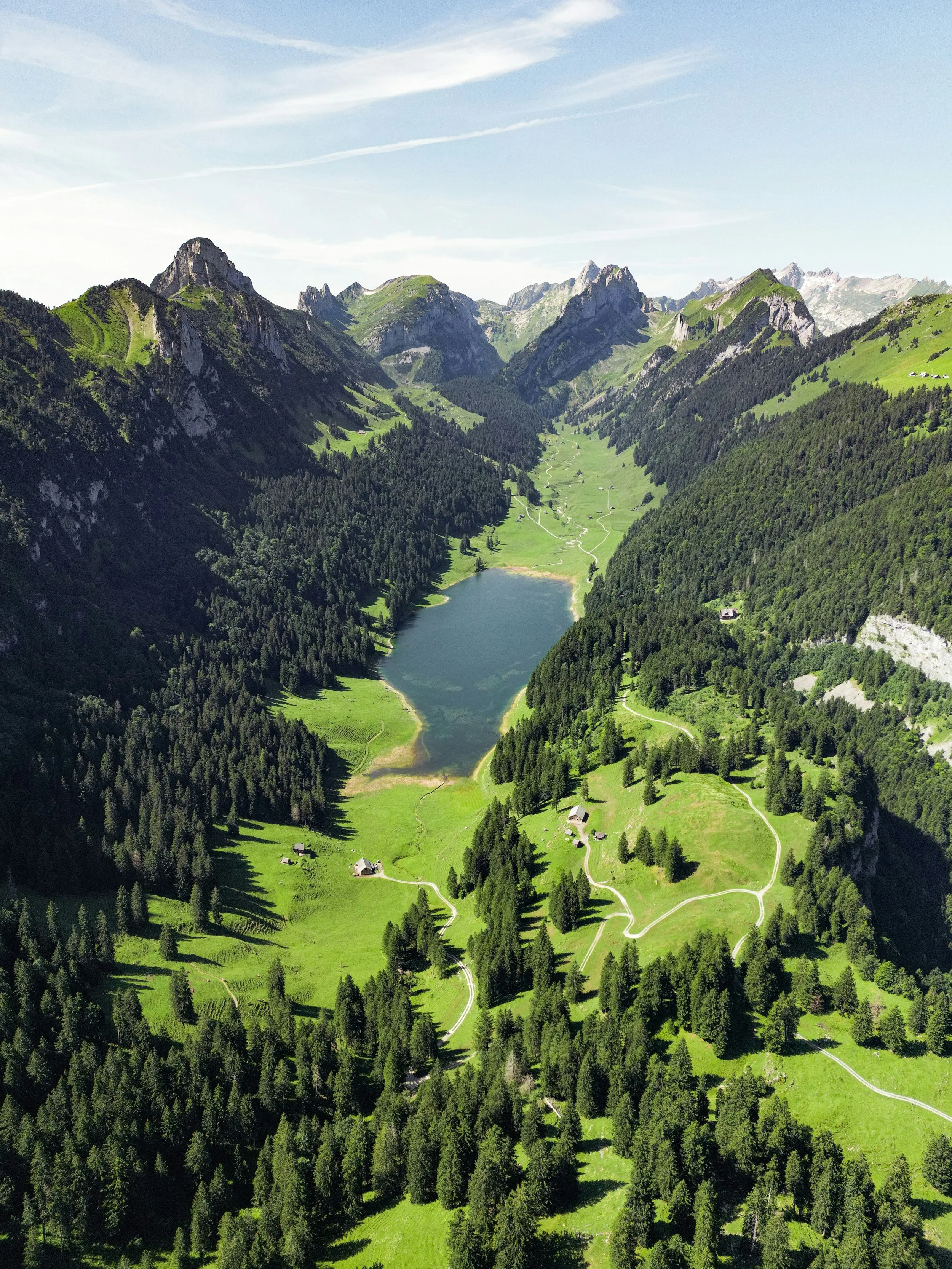 Vue aérienne d'une vallée alpine avec un lac au centre, entouré de montagnes et de forêts, avec des sentiers sillonnant la zone.