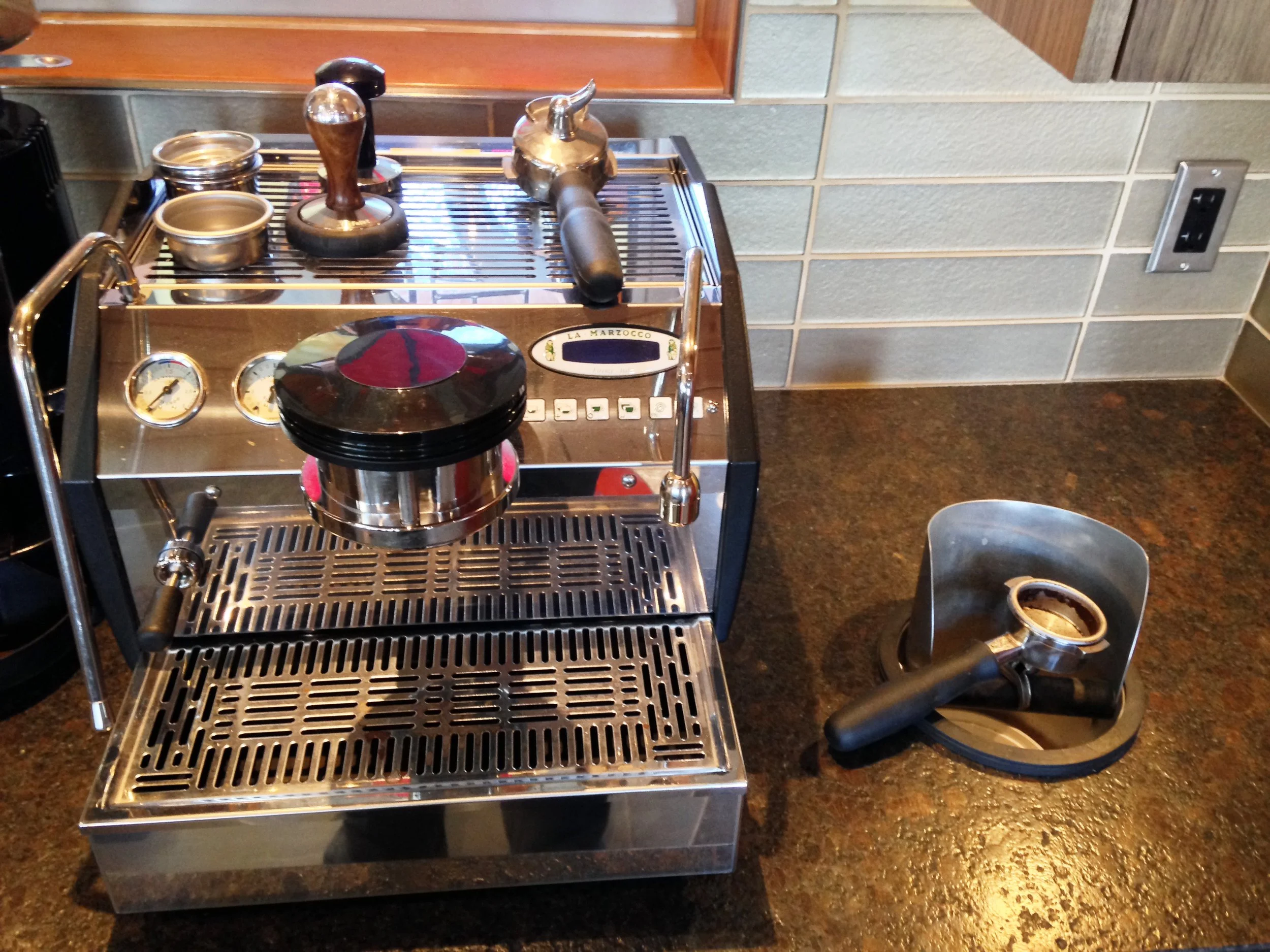 A commercial espresso machine with portafilters, tamper, and coffee cups on top, set on a brown granite countertop with tiled wall in background.