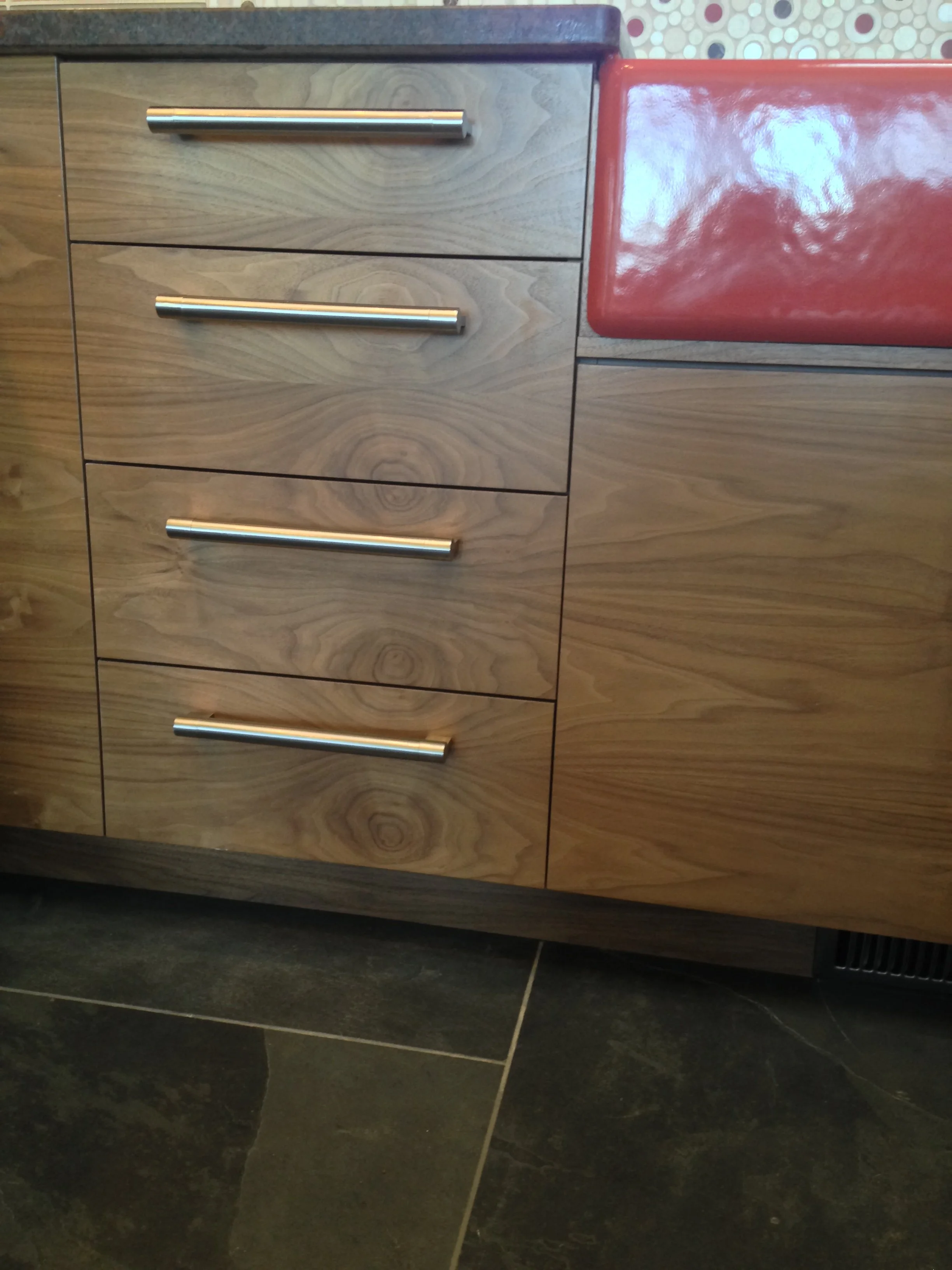 Close-up of a wooden kitchen cabinet with four drawers, each with a silver handle, next to a red countertop and patterned backsplash. Black tiled floor.