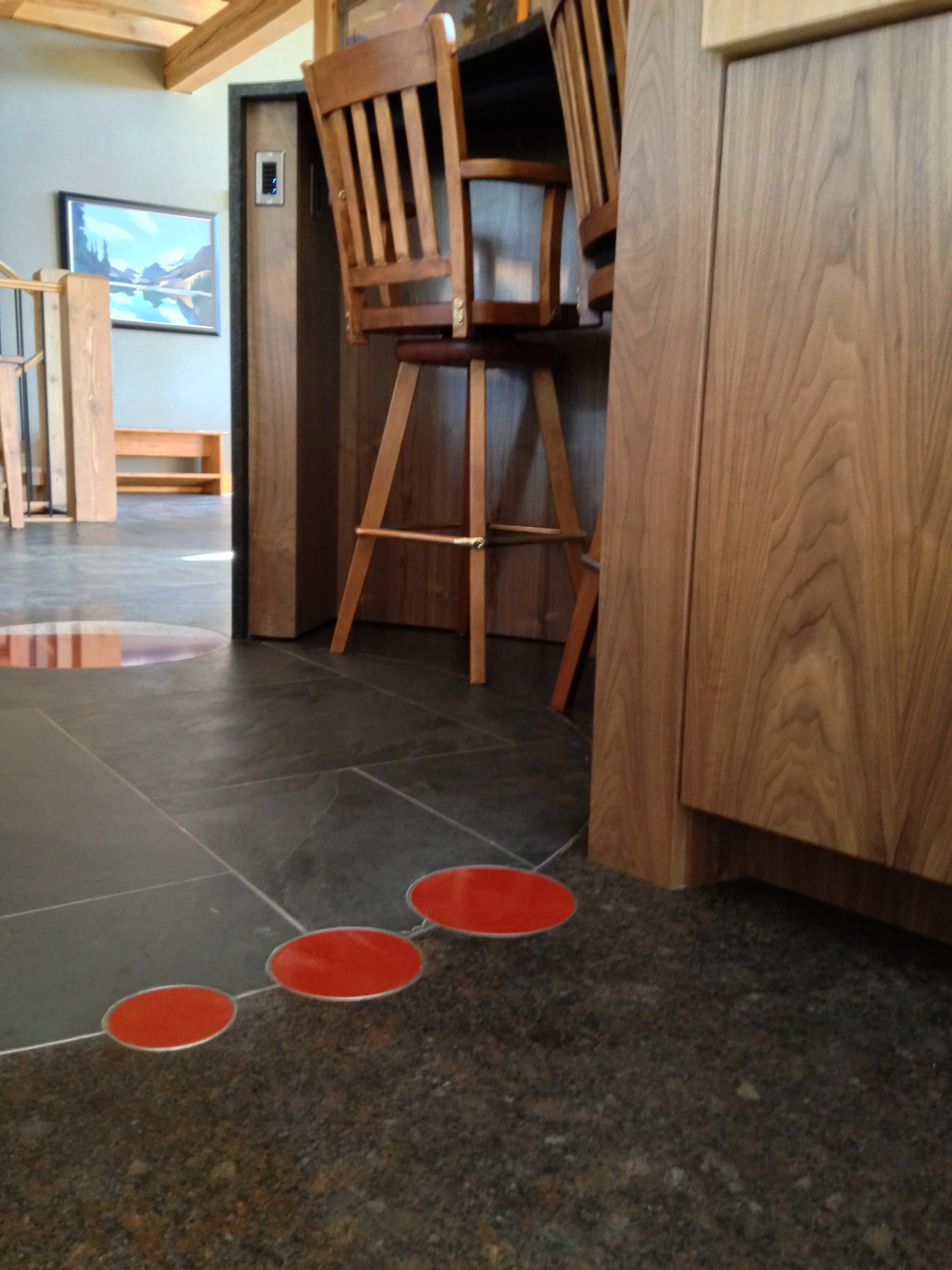 Close-up of a floor in a rustic indoor setting with three red circular sticker markers on the dark, textured tile floor. The background features wooden furniture and a framed landscape painting on a light green wall.