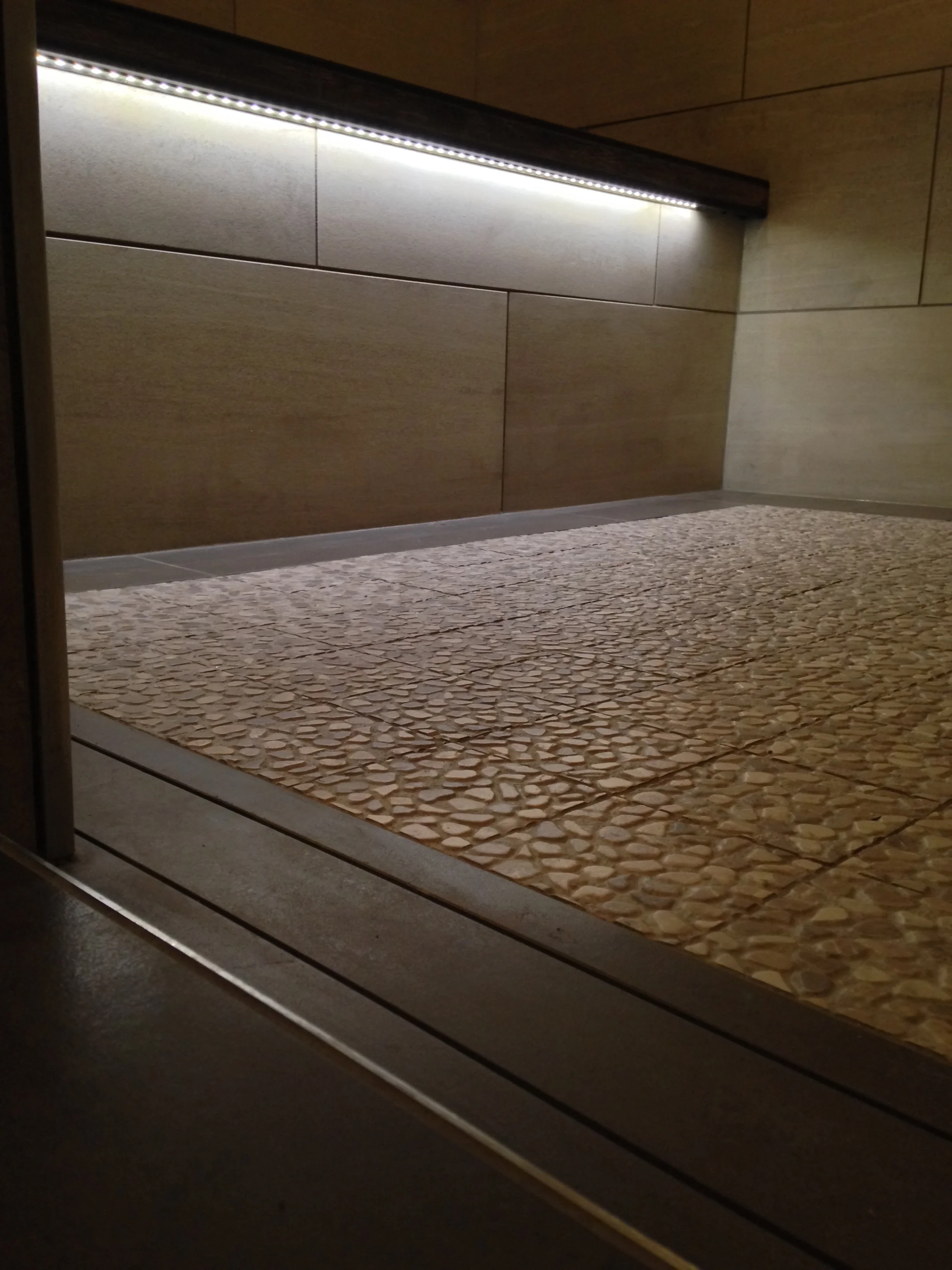 Close-up view of a tiled floor and wall inside an elevator, with a strip of LED lighting along the top edge of the wall.
