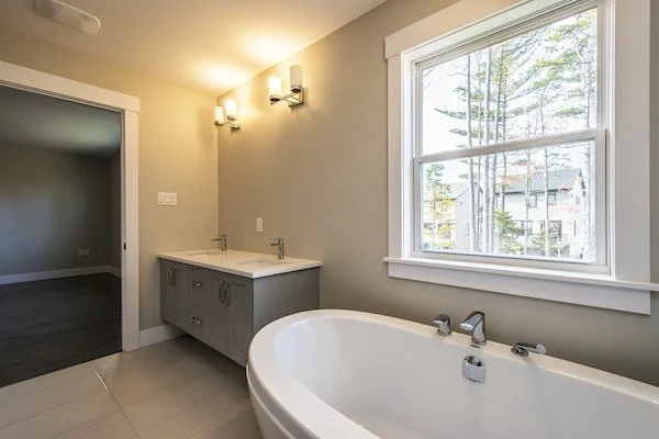 Bathroom with a large bathtub, a double sink vanity, and a window overlooking trees and neighboring houses.