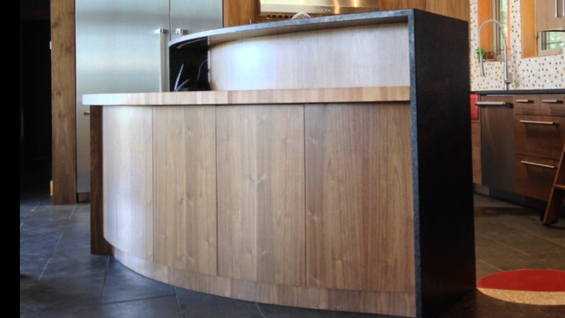 Kitchen island with wooden doors and a black counter with a curved rounded edge. In the background, there are brown wooden cabinets and a stainless steel refrigerator.